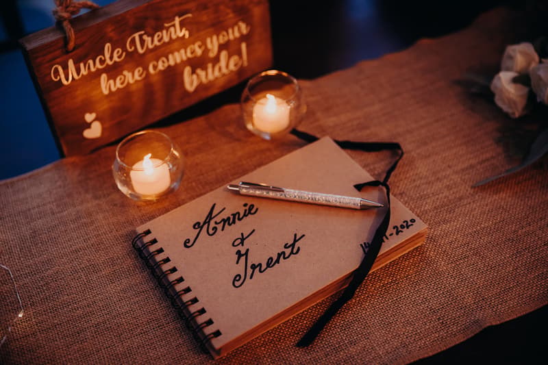 A wedding guest book with the names Annie and Trent written on the cover, placed on a burlap-covered table at Ocean View Estates. Two lit candles in glass holders and a wooden sign that reads 'Uncle Trent, here comes your bride!' are also visible.