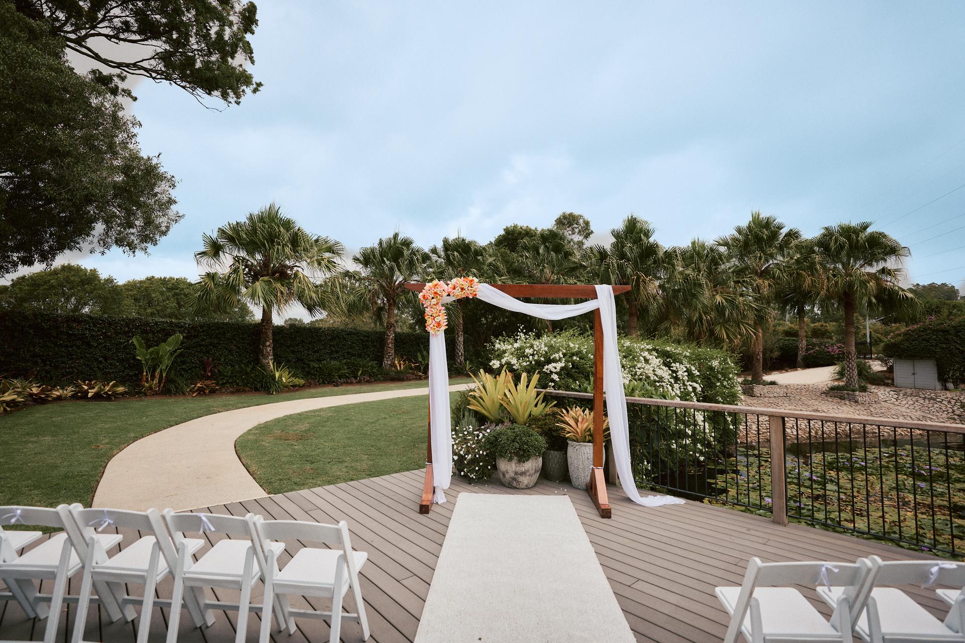 Empty outdoor wedding ceremony setup at Eatons Hill Hotel — Lakeside with a wooden arch decorated with white fabric and flowers, white chairs arranged on both sides of an aisle runner, surrounded by palm trees and greenery.