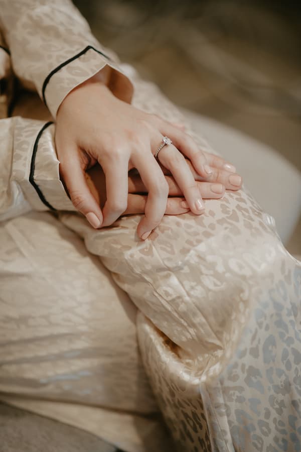 Close-up of a person's hands resting on their lap, wearing a light-colored satin garment with a subtle leopard print pattern and a ring with a prominent stone on the ring finger.