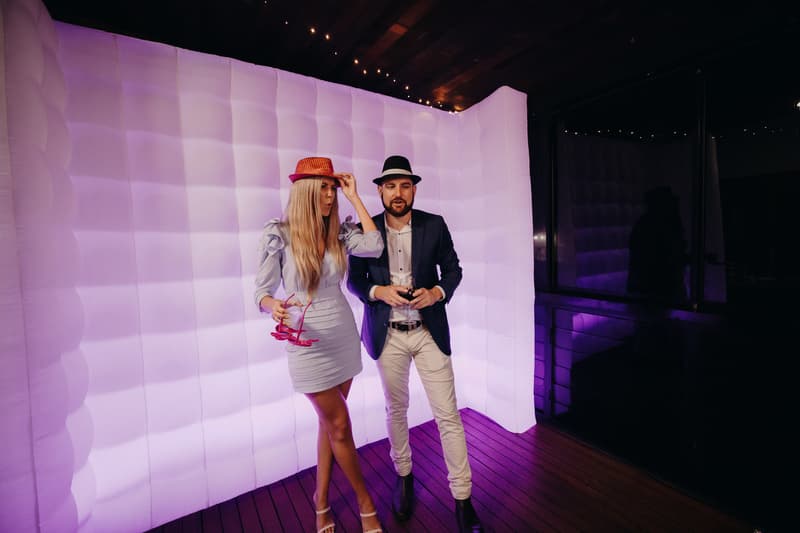 The bride Chelsea and the groom Brent stand in front of a white inflatable photo booth backdrop at Preston Peak Winery Functions Centre, each wearing a hat and holding props.