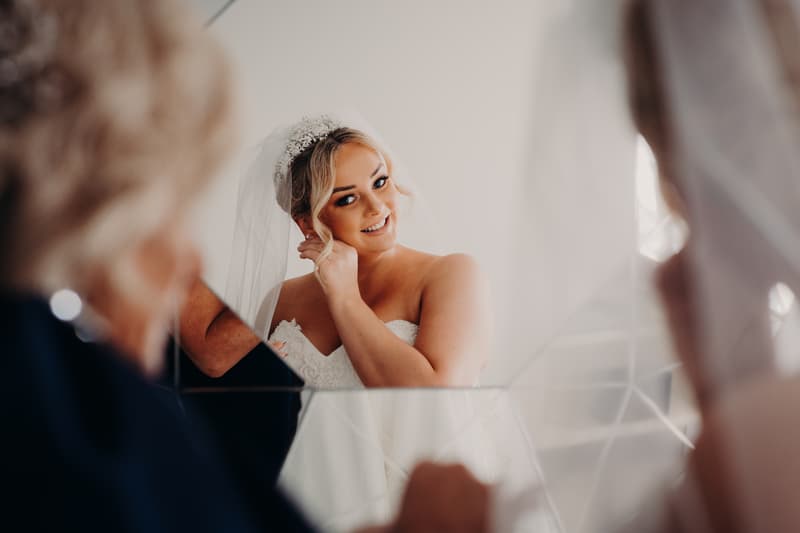 The bride Emily adjusts her earring while looking into a mirror, with an older woman partially visible in the foreground.