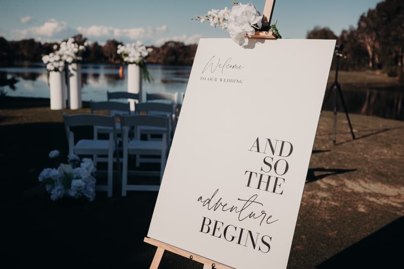 A wedding welcome sign reading 'And so the adventure begins' is displayed on an easel outdoors near a water body at The Tides — The Water's Edge, with white chairs and floral arrangements visible in the background.