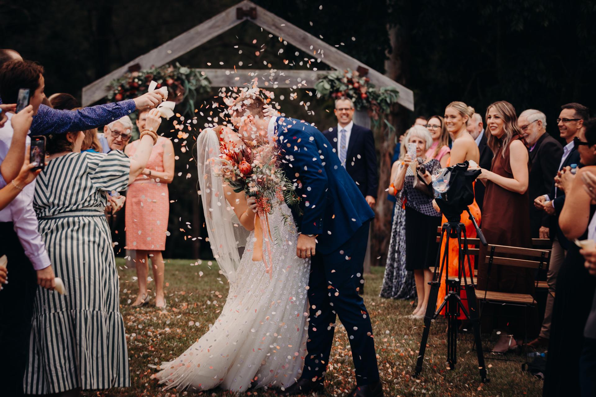 Bride Lilly and groom Connor kiss under a wooden arch decorated with flowers at Yabbaloumba Retreat — By The River as guests throw flower petals around them during the ceremony.