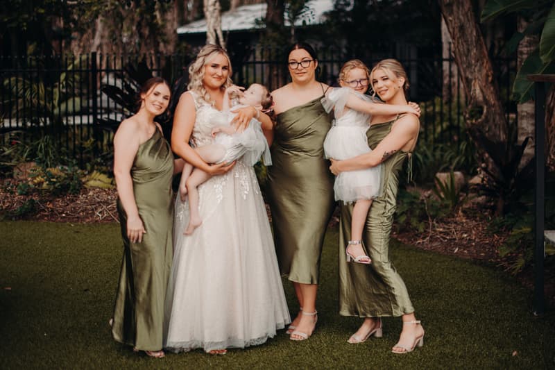 The bride Chloe stands with four bridesmaids dressed in olive green gowns at Sandstone Point Hotel Pavilion. Two bridesmaids each hold a young flower girl in white dresses.