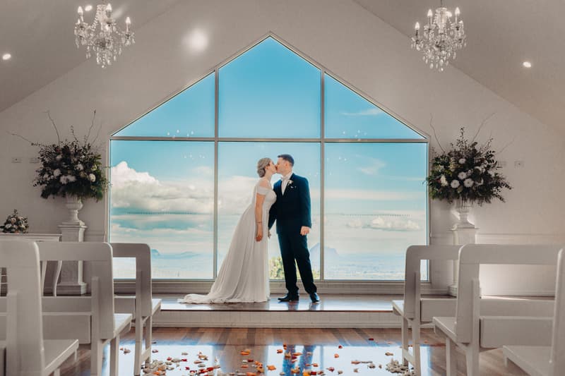Courtney and Liam kiss in front of a large triangular window at Tiffany's Maleny chapel with floral arrangements on either side and white pews in the foreground.