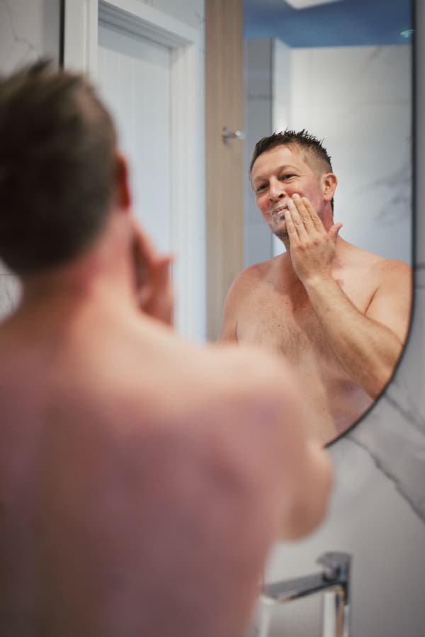 The groom Dane is shirtless and applying shaving cream to his face while looking into a bathroom mirror.