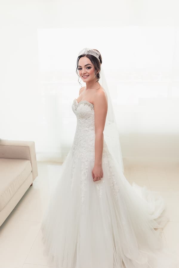 The bride Maryam stands indoors wearing a strapless white wedding gown with lace details and a tiara, next to a beige sofa.