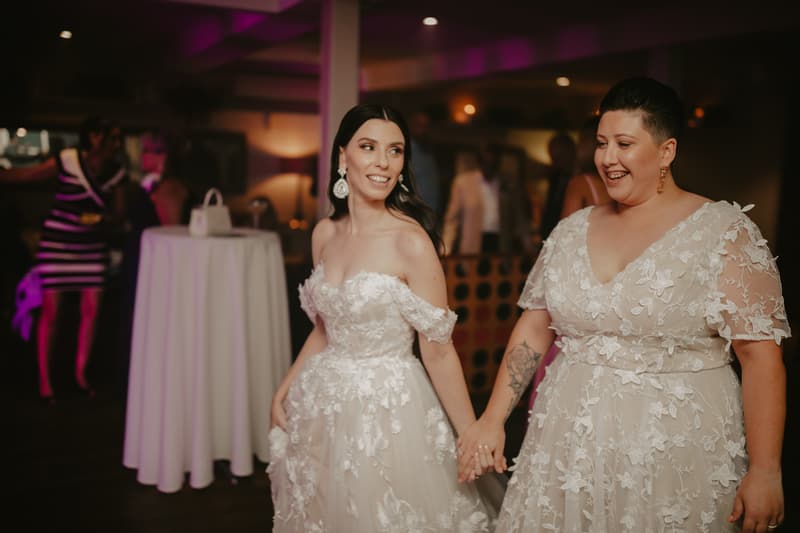Two brides in floral lace wedding dresses hold hands and smile at each other indoors with guests and a cocktail table in the background.
