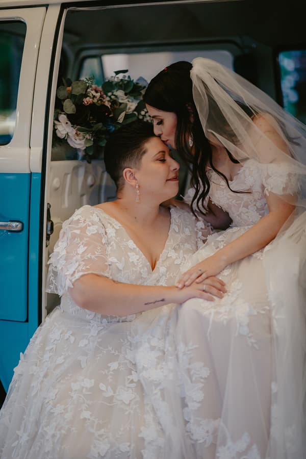 Two brides in white lace wedding dresses share an intimate moment inside a blue and white vintage vehicle, one bride sitting and the other leaning in to kiss her forehead, with a bouquet of flowers visible in the background.