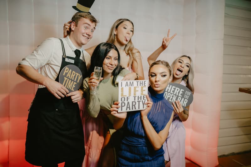 Five guests pose in front of a white inflatable photo booth backdrop at a party. One man wears a black apron and a small top hat prop, holding a sign that says 'PARTY TIME'. Four women surround him, two holding signs that read 'I AM THE LIFE OF THE PARTY' and 'HERE FOR THE OPEN BAR'. One woman holds a glass of champagne. They make playful facial expressions and hand gestures.
