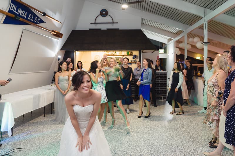 The bride Wing prepares to toss her bouquet to a group of female guests at the reception held at Bilinga Beach Weddings — Bilinga SLSC, with guests watching and smiling.