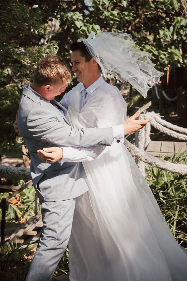 Jheng and Dane embrace outdoors at Sandstone Point Hotel during a couple portrait session, with Jheng wearing a wedding dress and veil blowing in the wind and Dane in a light grey suit.