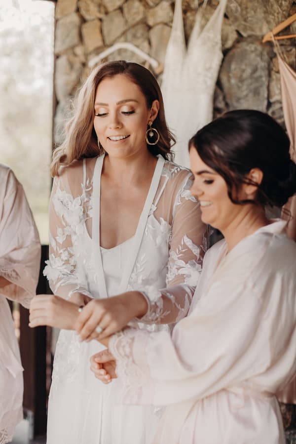 The bride Chelsea, wearing a white lace robe, is assisted by a bridesmaid in a light pink robe while preparing at Preston Peak Winery Functions Centre. Wedding dresses are hanging on the stone wall behind them.