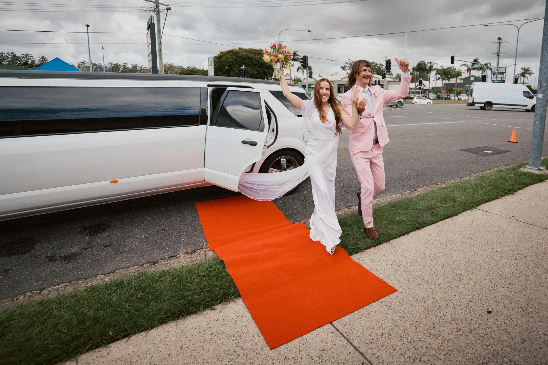 The bride Olivia and groom Jake step out of a white limousine onto a red carpet at White Horse Ranch, both raising their arms in celebration.