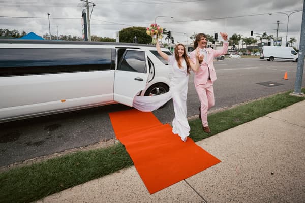 The bride Olivia and groom Jake step out of a white limousine onto a red carpet at White Horse Ranch, both raising their arms in celebration.