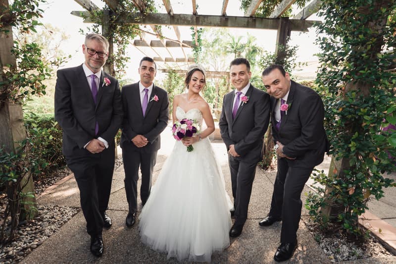 The bride Maryam stands holding a bouquet in the center under a pergola at Hillstone St Lucia — The Quartyard, flanked by four men in dark suits with purple ties and pink boutonnieres, likely the groom Pasha and groomsmen.