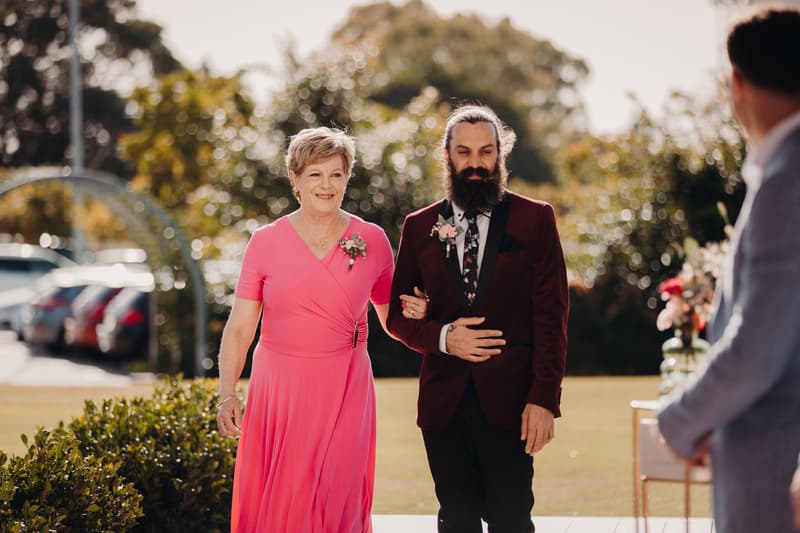 The groom walks arm-in-arm with an older woman, likely his mother, down the aisle at Sandstone Point Hotel — Pavilion during the ceremony.