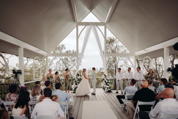 Bride Chloe and groom Brodie stand facing each other holding hands at the altar during their wedding ceremony at Sandstone Point Hotel — Pavilion, surrounded by bridesmaids in olive dresses and groomsmen in white shirts and light pants, with guests seated watching.