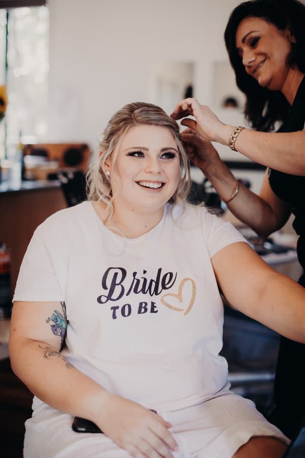 The bride Annie sits smiling while a woman styles her hair indoors at Ocean View Estates.
