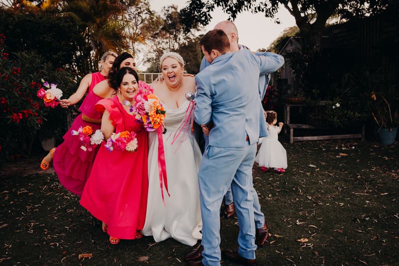 The bride Jacquelyne in a white gown and groom Arran in a light blue suit embrace and laugh with three bridesmaids in bright pink dresses holding colorful bouquets at Sandstone Point Hotel. A young flower girl in a white dress with pink shoes stands nearby on the grass.