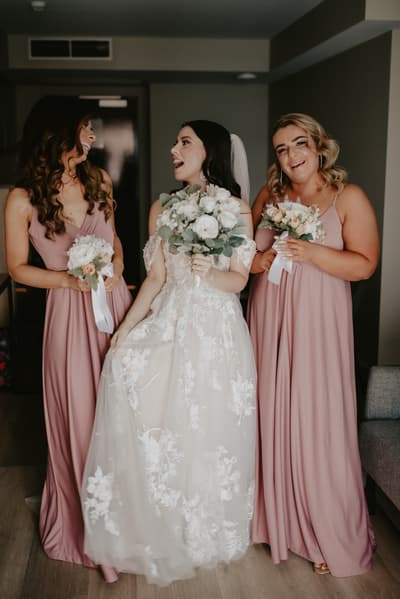 A bride in a white lace wedding gown and veil stands indoors between two bridesmaids wearing matching mauve dresses, each holding a bouquet of flowers. Best Brisbane wedding photographer by She Said Yes.