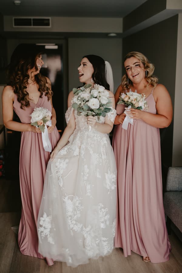 A bride in a white lace wedding gown and veil stands indoors between two bridesmaids wearing matching mauve dresses, each holding a bouquet of flowers.