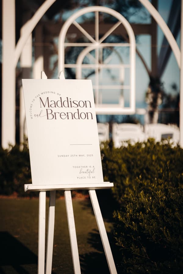 Wedding welcome sign for Madison and Brendon displayed on an easel at Sandstone Point Hotel — Pavilion, with white chairs and decorative arches in the background.