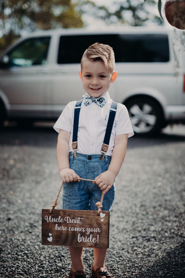 A young boy dressed in a white shirt, blue suspenders, blue shorts, and a floral bow tie stands outdoors on gravel holding a wooden sign that reads 'Uncle Trent, here comes your bride!' A white van is parked in the background.