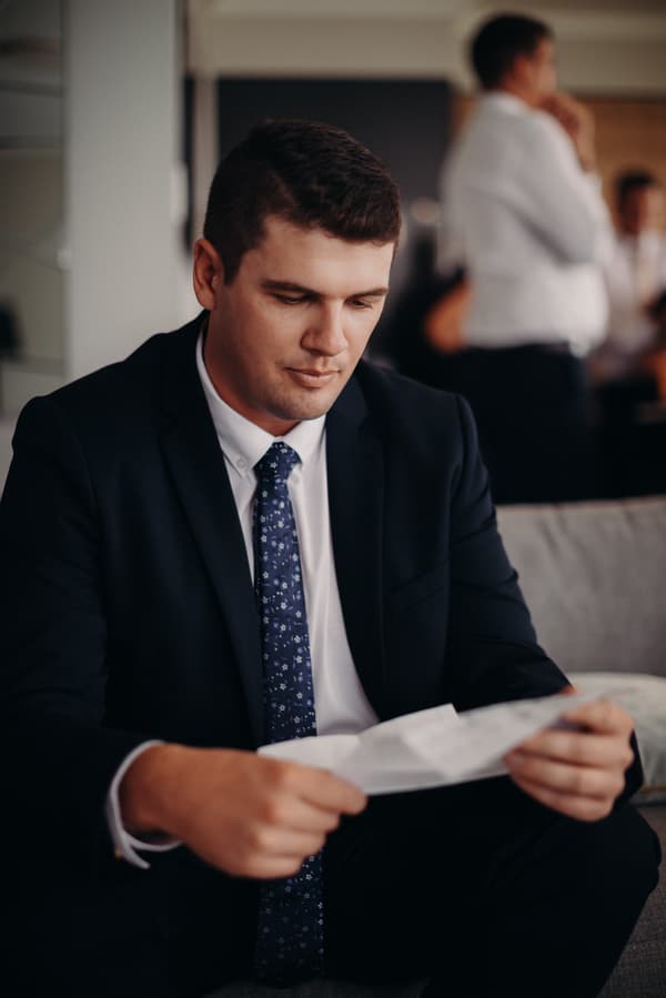 The groom Brent, dressed in a dark suit and patterned tie, is seated on a couch reading a piece of paper indoors at Preston Peak Winery Functions Centre. Other guests are visible blurred in the background.