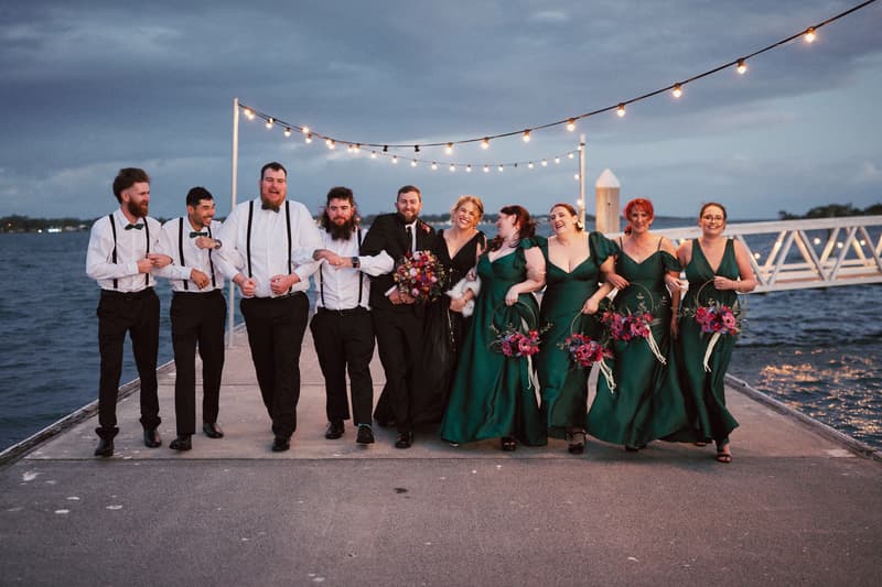 Christopher and Jayme with their bridal party stand linked arm-in-arm on a pier at dusk with string lights overhead and water in the background.