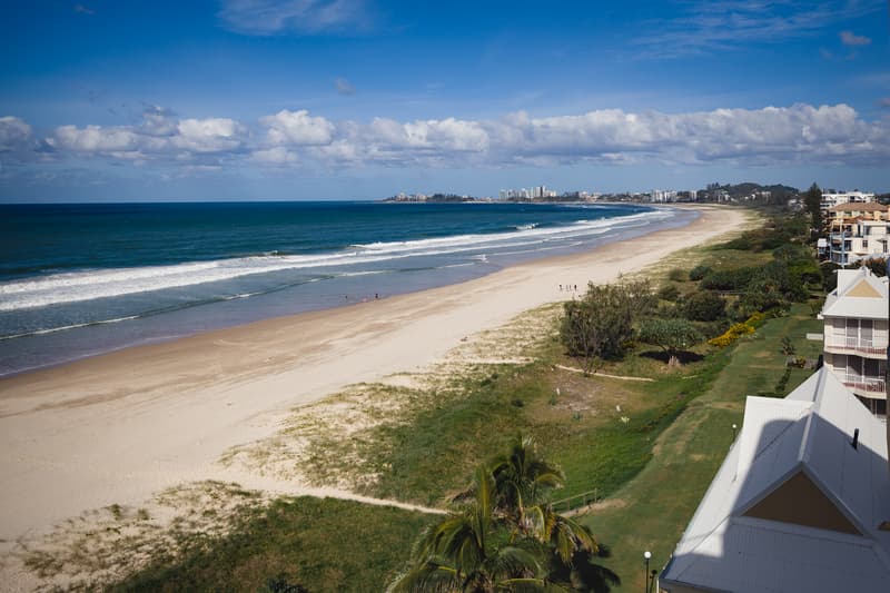 View of Bilinga Beach with sandy shore, ocean waves, scattered people on the beach, and buildings along the right side under a partly cloudy blue sky.