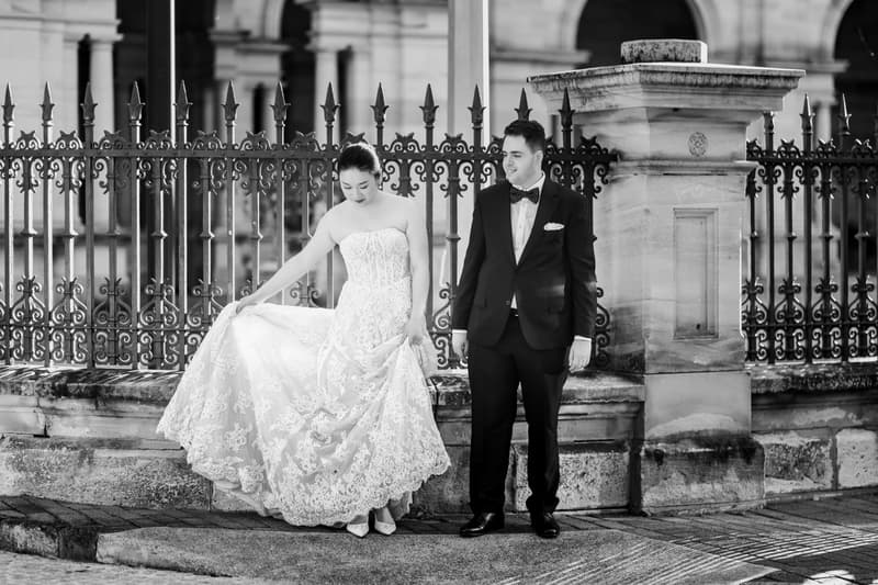 Aria in her wedding gown and Antony in a tuxedo stand near an ornate iron fence and stone pillar at Queensland Parliament building.