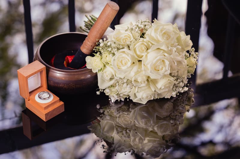 A bridal bouquet of white roses and baby's breath lies next to a wooden ring box containing a wedding ring and a decorative bowl with a wooden mallet on a reflective black surface at Toowong Rowing Club — The Malouf Room.
