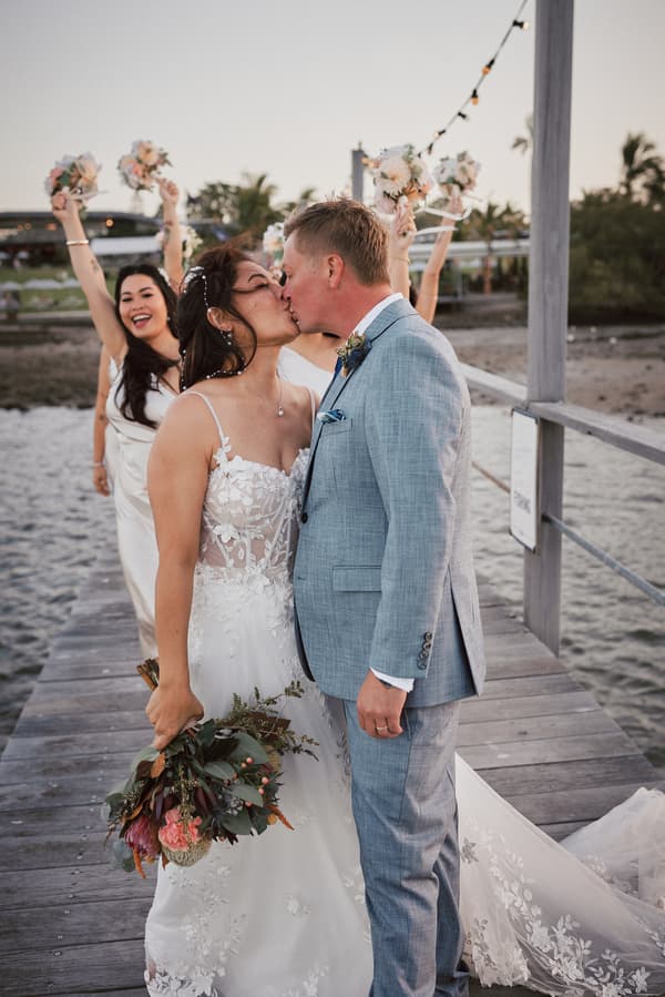 Bride Jheng and groom Dane kiss on a wooden pier at Sandstone Point Hotel, with bridesmaids behind them holding bouquets up in celebration.