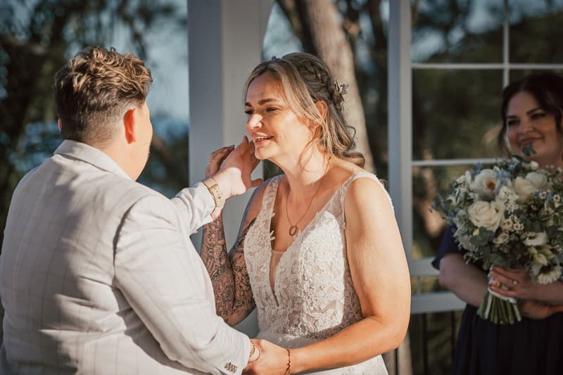 Brides Mandy and Selina share a moment during their wedding ceremony at Sandstone Point Hotel — The Pavilion, with a bridesmaid holding a bouquet in the background.