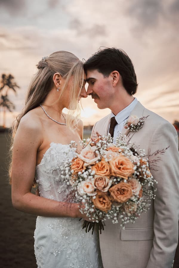 Morgan and Joel stand close together outdoors at Sandstone Point Hotel, with Morgan holding a bouquet of peach and cream roses and baby's breath.