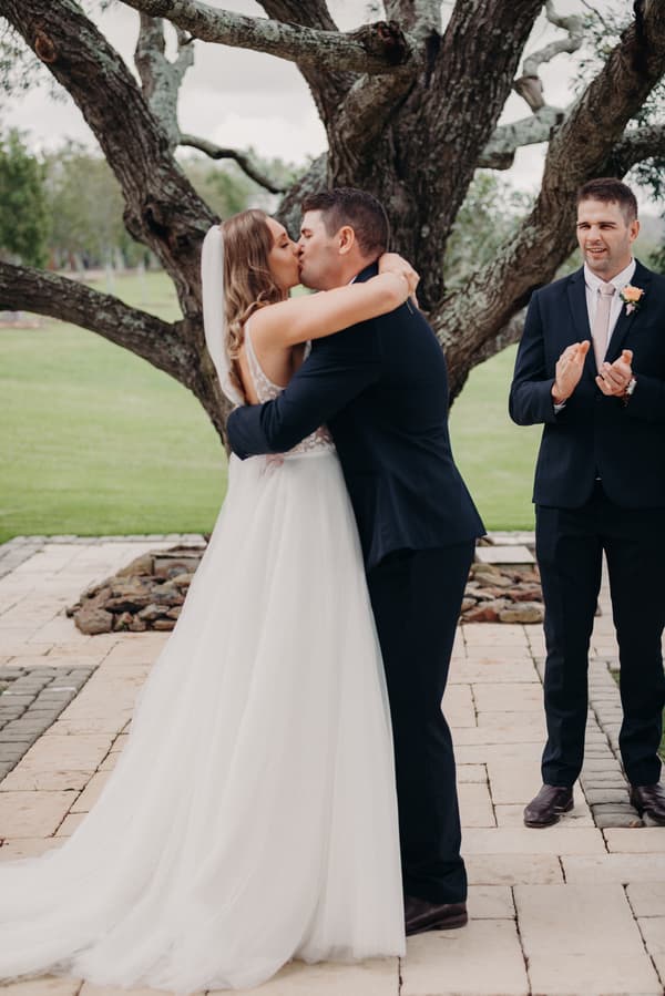 Chelsea and Brent share a kiss during their wedding ceremony at Preston Peak Winery Functions Centre, with a groomsman clapping nearby.
