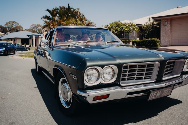 A group of people sit inside a dark green vintage car parked on a residential street near Ocean View Estates.