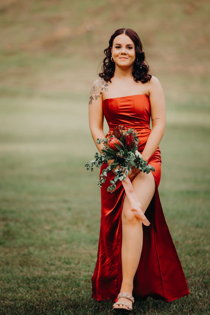 A bridesmaid in a red strapless dress holding a bouquet stands on grass. Wedding family photos by She Said Yes.