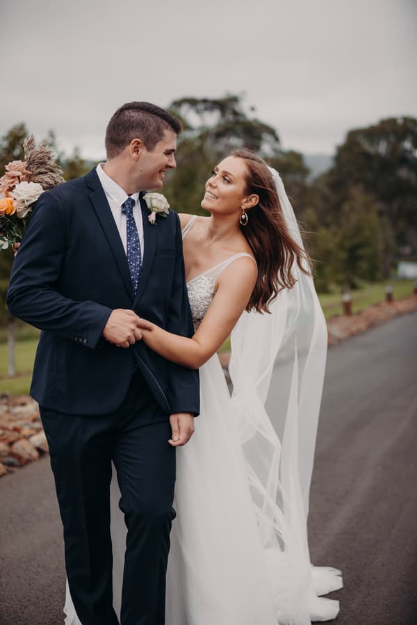 Chelsea the bride and Brent the groom walk arm in arm outdoors at Preston Peak Winery Functions Centre, with Chelsea holding a bouquet and wearing a veil.