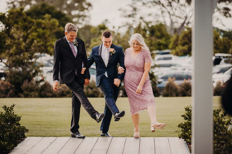 Groom Dylan links arms with an older man and woman, likely his parents, as they step forward together on the ceremony stage at Sandstone Point Hotel — Pavilion.