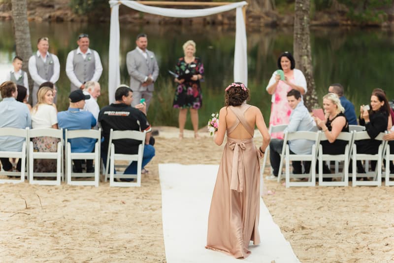 A bridesmaid walks down the aisle holding a bouquet during the wedding ceremony at Novotel Sunshine Coast Resort, with guests seated on either side and the wedding party and officiant standing near the altar.