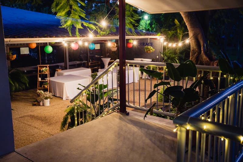 View of the decorated outdoor reception area at TOOWONG ROWING CLUB — The Malouf Room, featuring string lights, colorful paper lanterns, white-covered tables, potted plants, and a stair railing wrapped with fairy lights.