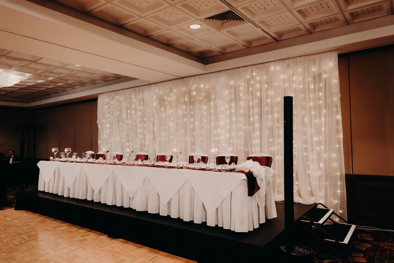 Empty head table set with glassware and white tablecloths on a raised platform with a backdrop of white drapes and string lights at Royal on the Park.