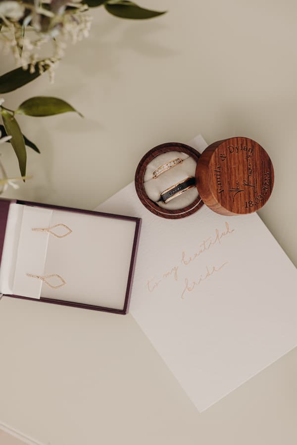 Wedding rings in a wooden box engraved with 'Emik & Dylan 18.03.2023', a pair of gold earrings in a box, and a handwritten note addressed 'to my beautiful bride' on a table.