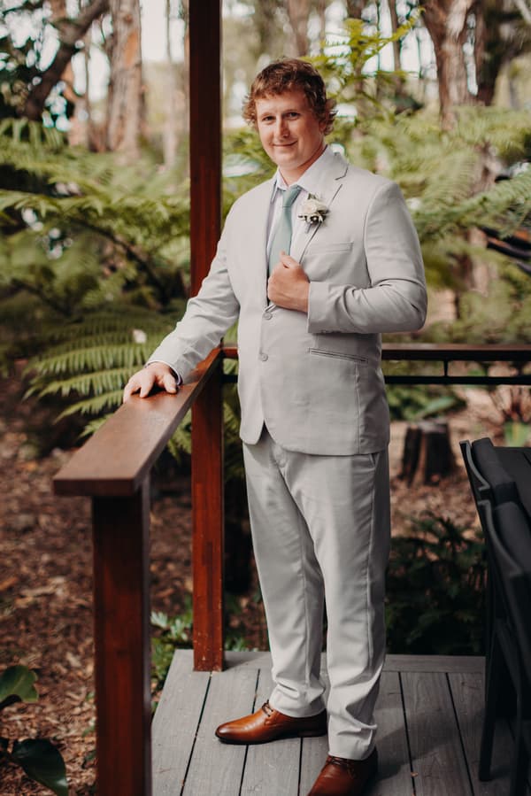 Brodie, the groom, poses alone on a wooden deck surrounded by greenery at Sandstone Point Hotel.