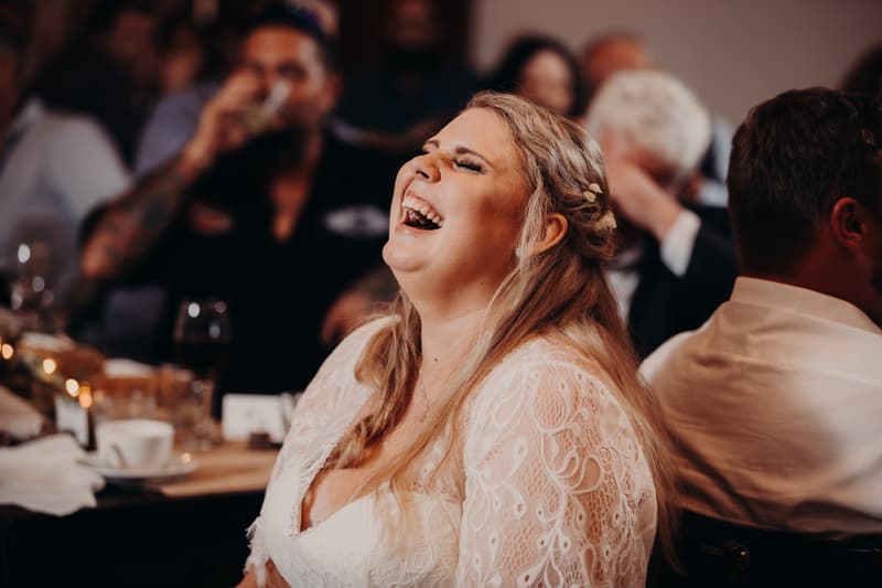 The bride Annie laughs joyfully while seated at a table during the reception at Ocean View Estates, with guests visible in the background.