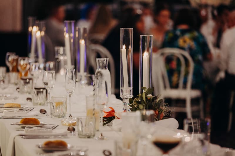 Reception table set with candles, glassware, cutlery, and bread rolls at Sandstone Point Hotel — Pumicestone Room, with guests seated in the background.