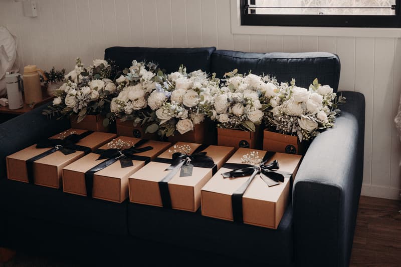 A dark blue couch at Sandstone Point Hotel Pavilion holds six brown gift boxes tied with black ribbons and six bouquets of white flowers arranged behind the boxes.