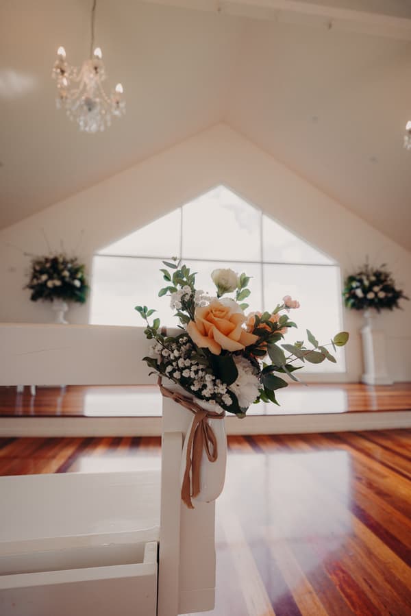Floral arrangement with peach and white flowers tied to a white wooden pew inside Tiffany's Maleny chapel, with large windows and chandeliers visible in the background.